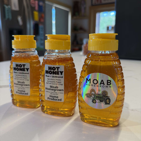 Three bottles of hot honey with yellow caps lined up on a white countertop in a kitchen setting. The labels show 'Hot Honey' and 'Moab Homegrown'.